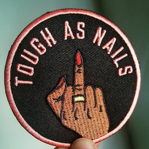 May include: A black and red embroidered patch with the text "TOUGH AS NAILS" and a stylized image of a hand giving the middle finger with a gold ring on the middle finger.