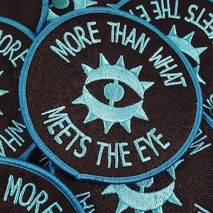 May include: A stack of black fabric patches with a blue border. The patches feature a stylized eye with a sunburst design and the text "More Than What Meets The Eye".