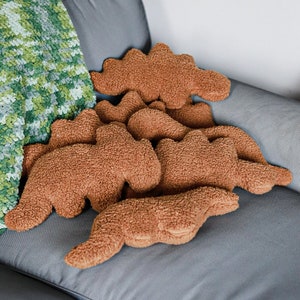 Pode incluir: Uma coleção de almofadas de pelúcia marrons em forma de dinossauro dispostas em um sofá cinza. As almofadas têm uma aparência macia e texturizada. Um cobertor de malha verde e branco é parcialmente visível no lado esquerdo do sofá.
