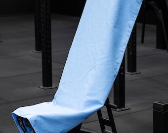 Può includere: Un asciugamano da palestra blu chiaro è drappeggiato su una panca per pesi nera in una palestra. L'asciugamano ha stampate le parole "Gym Towel".