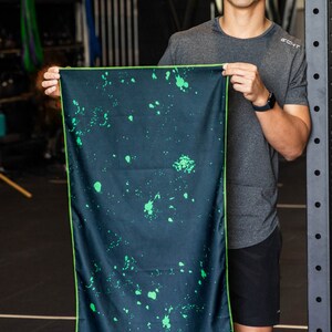 May include: A dark blue microfiber towel with a green splatter pattern and a lime green border. The towel is being held by a person in a grey shirt.
