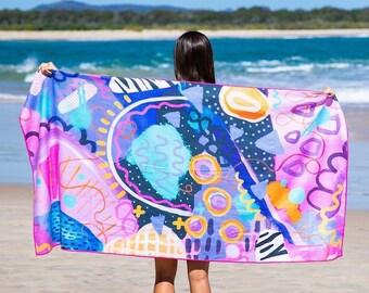 Può includere: Un asciugamano da spiaggia colorato con forme e motivi astratti in tonalità di rosa, blu, giallo e arancione. L'asciugamano è steso su una spiaggia sabbiosa.