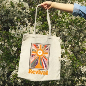 May include: A white canvas tote bag with a retro-style graphic of a sunburst with a cross in the center. The text "Revival" is printed in orange below the graphic.