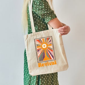 May include: A beige tote bag with a colorful retro design featuring a cross and the word "Revival".