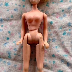 May include: A vintage doll with blonde hair and blue eyes. The doll is made of plastic and has articulated joints. The doll is wearing no clothing and is in a standing position. The doll is on a patterned fabric.