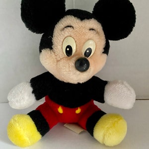 May include: A plush Mickey Mouse toy with black ears, a black body, red shorts with yellow buttons, and yellow shoes. The toy has a white face with black eyes and a black nose.