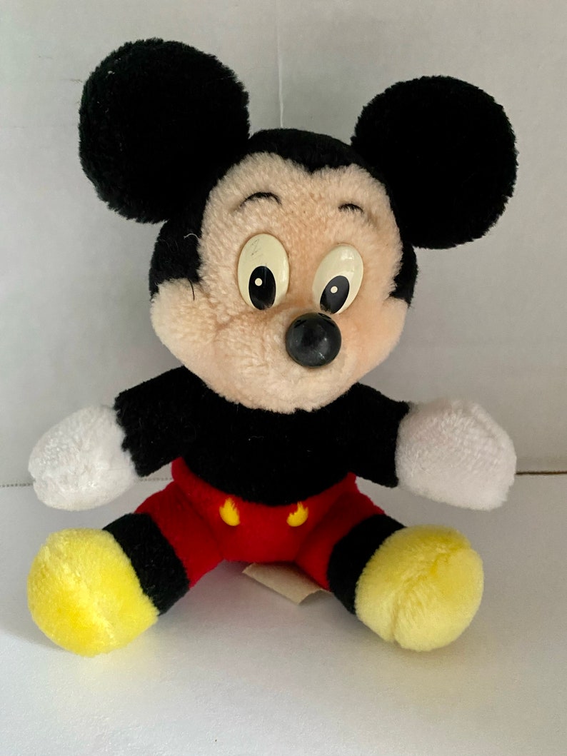 May include: A plush Mickey Mouse toy with black ears, a black body, red shorts with yellow buttons, and yellow shoes. The toy has a white face with black eyes and a black nose.
