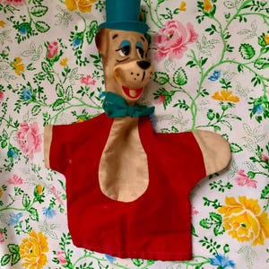 May include: A vintage hand puppet featuring a cartoon dog character. The puppet has a tan face with blue eyes, a black nose, and a red smiling mouth. It wears a teal top hat and bow tie, and a red shirt with tan accents.