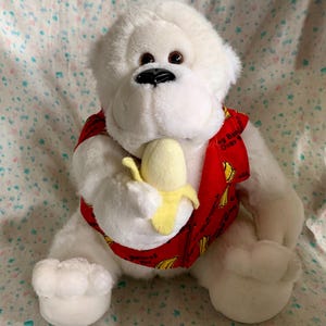 May include: A plush white gorilla toy holding a yellow banana. The gorilla wears a red vest with a banana pattern and the text "Going Bananas Over". The toy has brown eyes and a black nose.