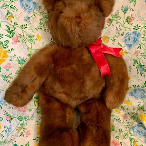 May include: A brown teddy bear with a red ribbon around its neck. The plush toy has a soft, furry texture and is lying on a floral patterned fabric with blue, yellow, and pink flowers.