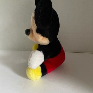 May include: A plush Mickey Mouse toy sitting with its legs crossed. The toy is black with red shorts and yellow shoes. The toy has a black nose and large black ears.