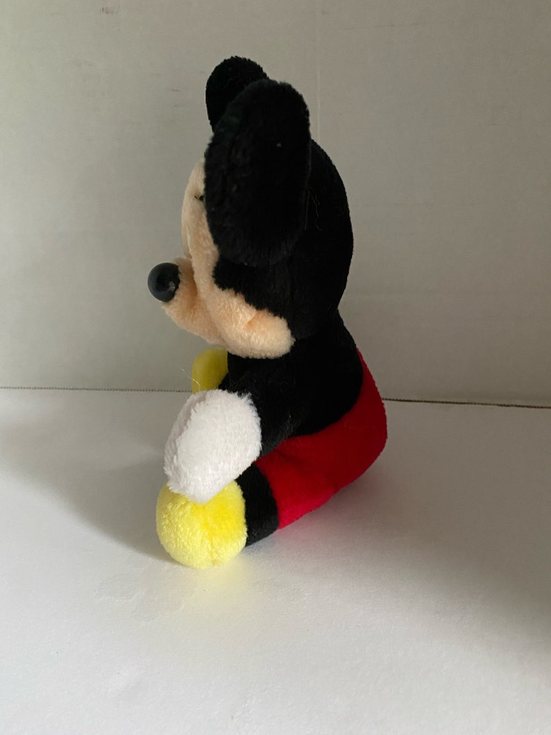 May include: A plush Mickey Mouse toy sitting with its legs crossed. The toy is black with red shorts and yellow shoes. The toy has a black nose and large black ears.