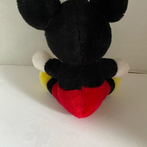 May include: A black and red plush Mickey Mouse toy sitting on a white surface. The toy has large black ears, a black body, white gloves, yellow shorts, and a red heart-shaped belly.
