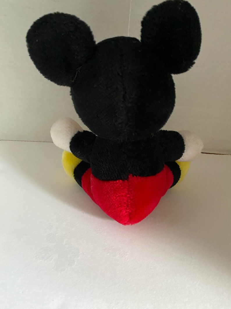 May include: A black and red plush Mickey Mouse toy sitting on a white surface. The toy has large black ears, a black body, white gloves, yellow shorts, and a red heart-shaped belly.