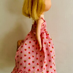 May include: A handmade doll with a light skin tone and blonde hair. The doll wears a pink dress patterned with red strawberries. The dress has a ruffled hem and straps. The doll's arms are visible.