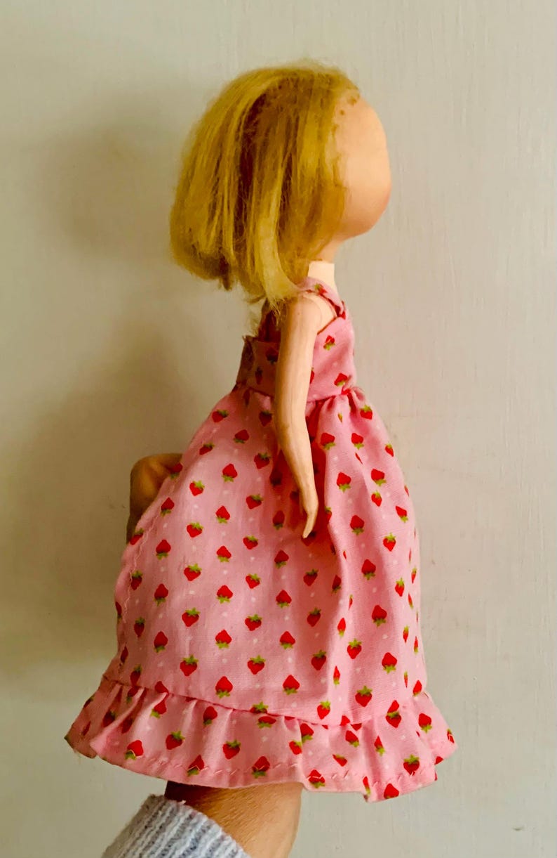 May include: A handmade doll with a light skin tone and blonde hair. The doll wears a pink dress patterned with red strawberries. The dress has a ruffled hem and straps. The doll's arms are visible.
