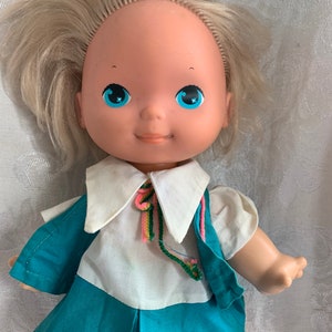 May include: A vintage doll with blonde hair, blue eyes, and a white and teal dress. The doll has a sweet expression and is wearing a colorful scarf.