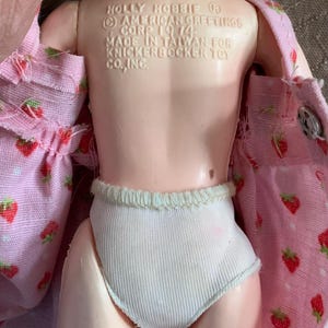 May include: A vintage Holly Hobbie doll with a pink strawberry-patterned dress and white ribbed underwear. The doll's back is visible, with text indicating it was made in Taiwan in 1974 by Knickerbocker Toy Co., Inc.
