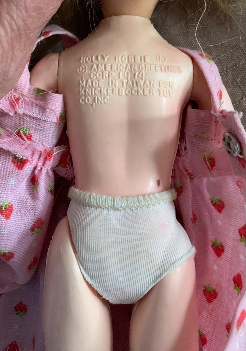 May include: A vintage Holly Hobbie doll with a pink strawberry-patterned dress and white ribbed underwear. The doll's back is visible, with text indicating it was made in Taiwan in 1974 by Knickerbocker Toy Co., Inc.