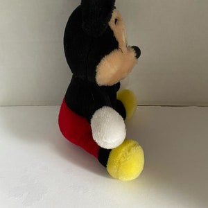 May include: A plush Mickey Mouse toy sitting with his legs crossed. He is wearing red shorts and yellow shoes. His ears are black and his face is tan.