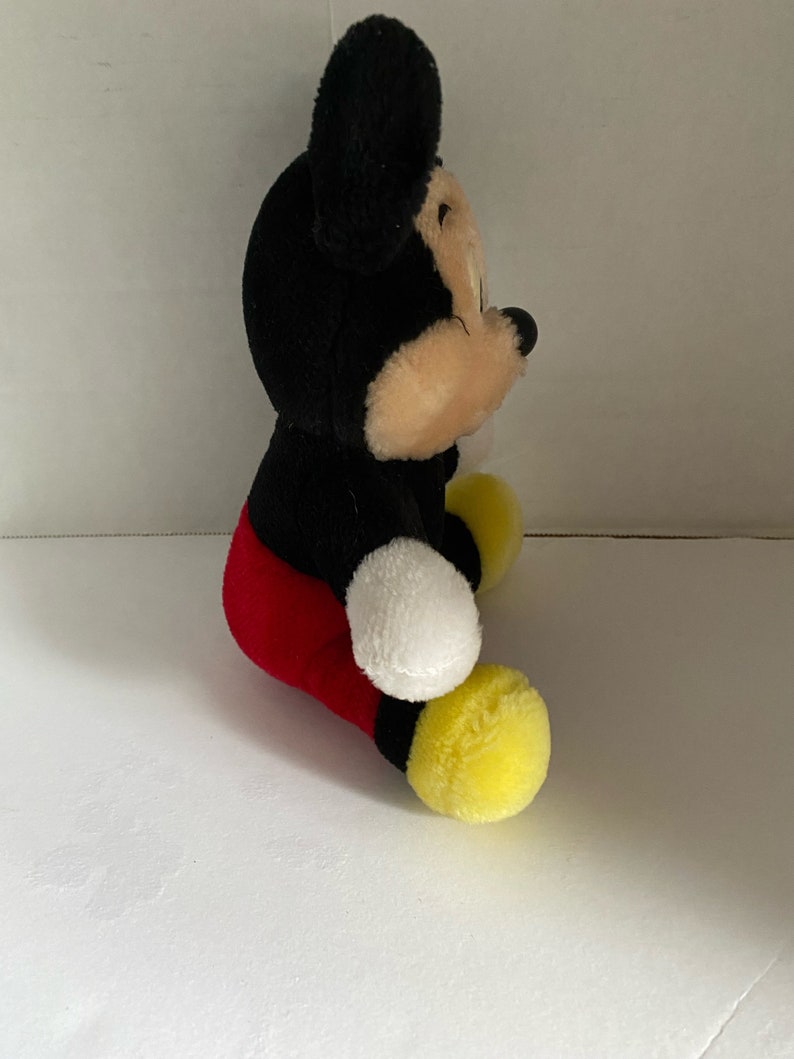 May include: A plush Mickey Mouse toy sitting with his legs crossed. He is wearing red shorts and yellow shoes. His ears are black and his face is tan.