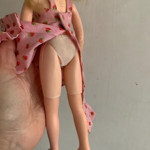 May include: A vintage doll with blonde hair, wearing a pink dress with a strawberry pattern. The doll's legs are visible, and it is held by a hand. The doll's dress is partially open.