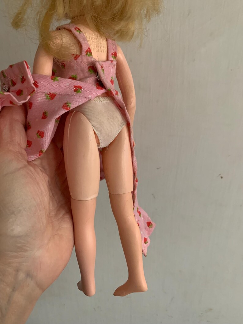 May include: A vintage doll with blonde hair, wearing a pink dress with a strawberry pattern. The doll's legs are visible, and it is held by a hand. The doll's dress is partially open.