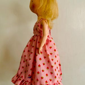May include: A vintage doll with blonde hair and a pink dress adorned with red strawberries. The dress has a ruffled hem and a simple, sleeveless design. The doll's face has painted features.
