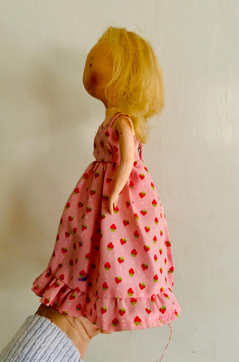 May include: A vintage doll with blonde hair and a pink dress adorned with red strawberries. The dress has a ruffled hem and a simple, sleeveless design. The doll's face has painted features.