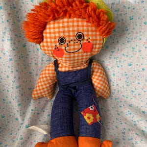 May include: A vintage handmade doll with an orange and white checked face and body, orange yarn hair, and a green hat. The doll wears blue denim dungarees and orange shoes. A small patch with a flower design is sewn on the leg.