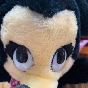 May include: Close-up of a plush toy character with a black head and ears, a light yellow face, and large, expressive eyes. A pink bow with white polka dots sits atop the head. The toy has a black nose and a pink and white polka dot bow around the neck.