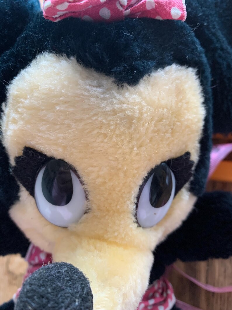 May include: Close-up of a plush toy character with a black head and ears, a light yellow face, and large, expressive eyes. A pink bow with white polka dots sits atop the head. The toy has a black nose and a pink and white polka dot bow around the neck.