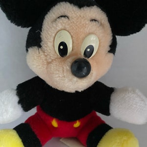 May include: A plush Mickey Mouse toy with black ears, a black nose, and yellow shoes. The toy is wearing red shorts with black trim and yellow buttons.