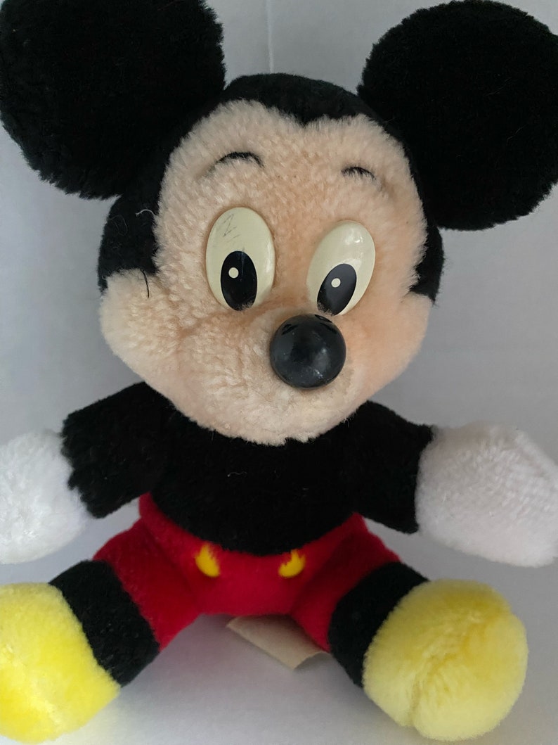 May include: A plush Mickey Mouse toy with black ears, a black nose, and yellow shoes. The toy is wearing red shorts with black trim and yellow buttons.