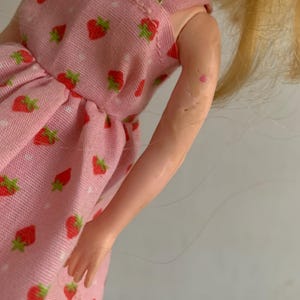 May include: A doll wearing a pink dress with a strawberry pattern. The dress has a white polka dot design and red strawberries with green leaves. The doll has blonde hair and a light skin tone.