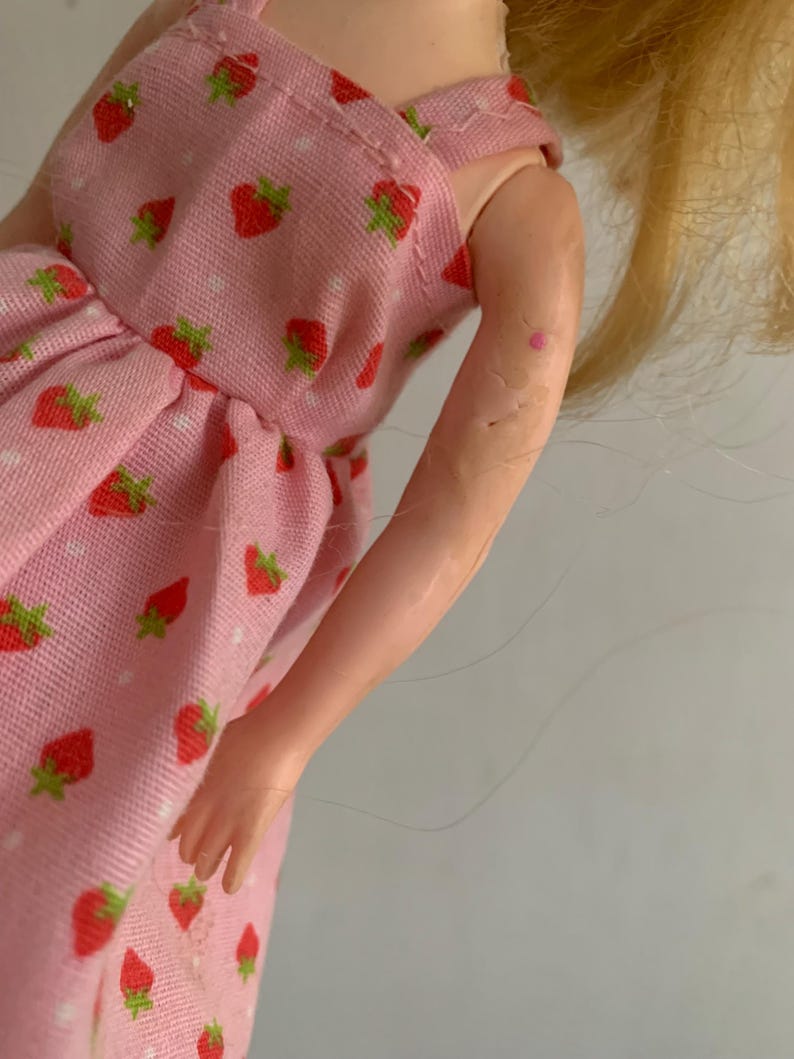 May include: A doll wearing a pink dress with a strawberry pattern. The dress has a white polka dot design and red strawberries with green leaves. The doll has blonde hair and a light skin tone.