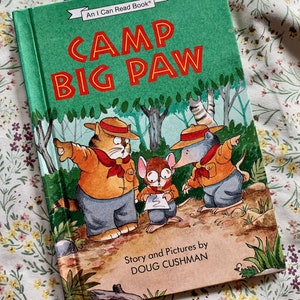May include: A green book cover with the title "Camp Big Paw" in red and white letters. The book is part of the "An I Can Read Book" series. The cover features an illustration of three animals in a forest setting: a cat, a mouse, and a mole. The text "Story and Pictures by DOUG CUSHMAN" is printed at the bottom of the cover.