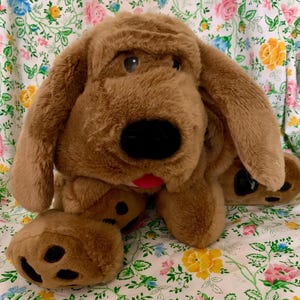 May include: A plush dog toy with soft brown fur, long floppy ears, and a large black nose. The stuffed animal features a small red tongue visible near the snout and dark brown paw pads on its feet, showcasing a classic design with a friendly expression.