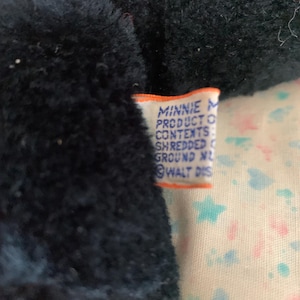 May include: Close-up of a black plush toy with a tag that reads "MINNIE PRODUCT CONTENTS SHREDDED GROUND NL WALT DIS". The toy is on a patterned fabric with stars and hearts.