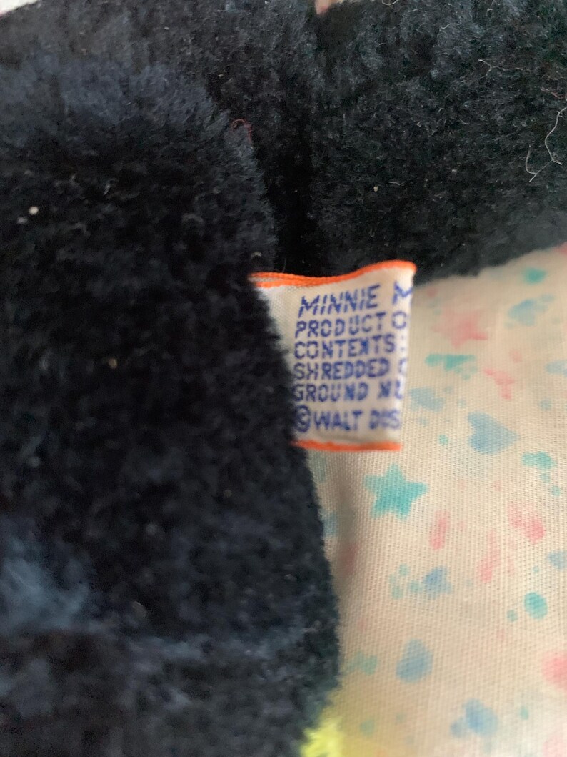 May include: Close-up of a black plush toy with a tag that reads "MINNIE PRODUCT CONTENTS SHREDDED GROUND NL WALT DIS". The toy is on a patterned fabric with stars and hearts.