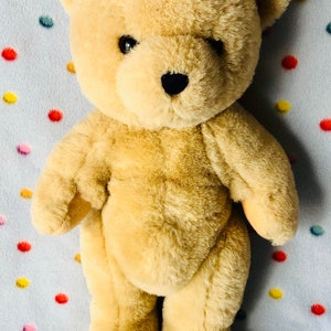 May include: A plush, tan teddy bear with a black nose and eyes stands on a light blue surface dotted with colorful circles. The bear has a soft, furry texture and a classic design, perfect for cuddling.