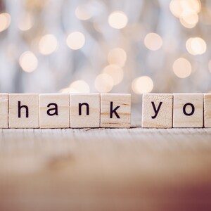 May include: The words "thank you" spelled out with wooden letter tiles against a blurred background of white and gold lights.