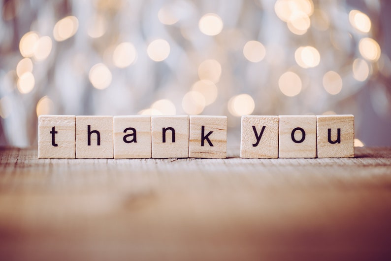 May include: The words "thank you" spelled out with wooden letter tiles against a blurred background of white and gold lights.