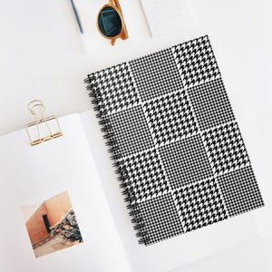 May include: A spiral-bound notebook with a black and white houndstooth pattern cover. The notebook is open, revealing blank white pages. A gold paper clip and sunglasses are also visible.