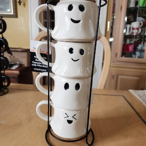 Könnte beinhalten: Ein Stapel von vier weißen Keramikbechern mit schwarzen Geistergesichtern, gehalten in einem schwarzen Metallgestell. Jeder Becher hat einen anderen Ausdruck. Die Becher sind Teil eines Halloween-Sets.