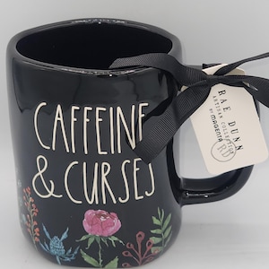 May include: A black ceramic mug featuring the white text "CAFFEINE & CURSES" and a colorful floral pattern at the base. A black ribbon is tied to the handle, holding a white tag that reads "RAE DUNN ARTISAN COLLECTION BY MAGENTA RD".