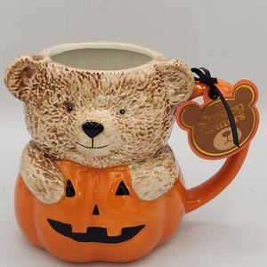 Peut inclure: Une tasse en céramique en forme d'ours en peluche brun sortant d'une citrouille orange sculptée en lanterne. L'ours a une fourrure texturée et la citrouille présente un visage noir. Une étiquette en forme d'ours indique Creepy Critters.