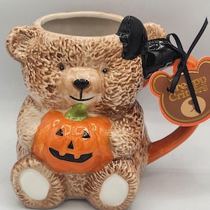 Könnte beinhalten: Ein Keramikbecher in Form eines braunen Teddybären, der einen orangefarbenen Kürbis hält. Der Bär hat eine schwarze Schleife auf dem Kopf und ein am Henkel befestigtes Etikett mit der goldenen Aufschrift Creepy Critters auf braunem Grund.
