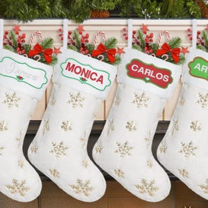 May include: Four white Christmas stockings with gold snowflake patterns and personalized name tags. Each stocking has a different name: James, Monica, Carlos, and Arturo. The stockings are decorated with red bows, candy canes, and greenery.