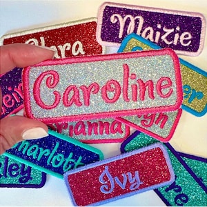 Könnte beinhalten: Eine Sammlung glitzernder Namensaufnäher in verschiedenen Farben, darunter Rosa, Rot, Lila, Blau und Gold. Jeder Aufnäher zeigt einen anderen Namen, der in einer Kontrastfarbe gestickt ist, wie z. B. "Caroline" in Pink auf silbernem Hintergrund.